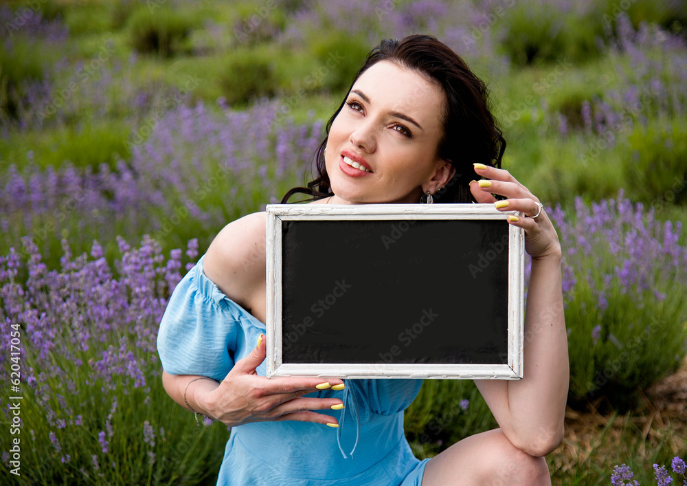 beautiful brunette girl in a blue dress sitting in a lavender field ,holding a wooden sign for inscriptions in her hands and looking up