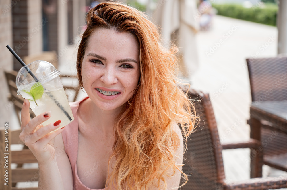 Young beautiful red-haired woman with braces drinks cooling lemonade outdoors in summer. Portrait of a smiling girl with freckles. Young beautiful red-haired woman with braces drinks cooling lemonade outdoors in summer. Portrait of a smiling girl with freckles.