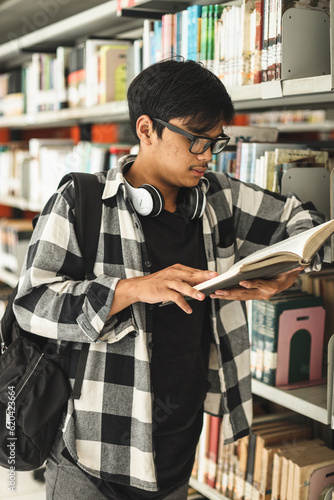Young teenager student wearing eyeglasses and having headphones on neck standing by bookcase and choosing books. Education concept