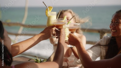 Multiracial women cheering and clinking glasses with cocktails in sea view rooftop cafe. Multiethnic female friends drink beverages have fun at cocktail party in a tropical outdoor restaurant.