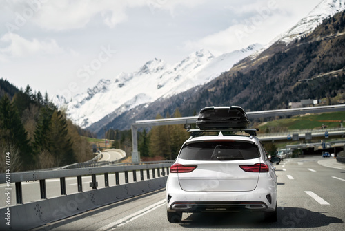 White wagon with roof box storage on the highway in Switzerland. Modern family car adventures in the Alpine Mountains in Europe. Plastic luggage compartment on a car roof. Road trip getaway concept. © AlexGo