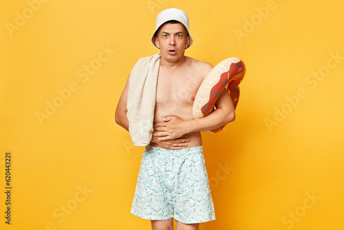 Shocked sick adult man wearing shorts swimsuit and panama holding donut rubber ring and bag isolated on yellow background suffering stomachache tropical food poisoning