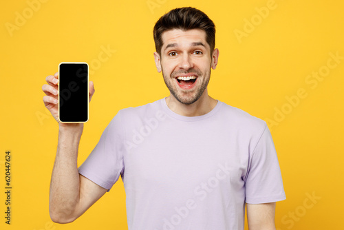 Young surprised man wear light purple t-shirt casual clothes hold in hand use mobile cell phone with blank screen workspace area isolated on plain yellow background studio portrait. Lifestyle concept.
