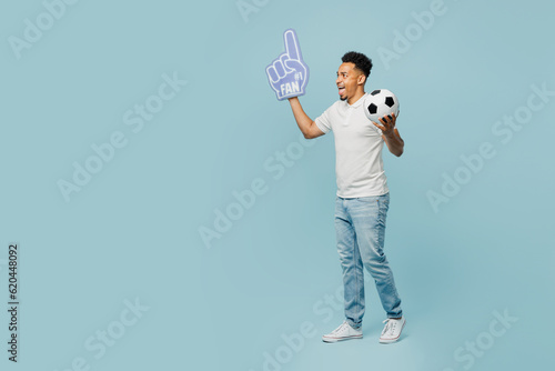 Full body side view young man fan wearing basic t-shirt foam 1 fan glove finger up cheer up support football sport team hold soccer ball watch tv live stream isolated on plain blue color background. © ViDi Studio