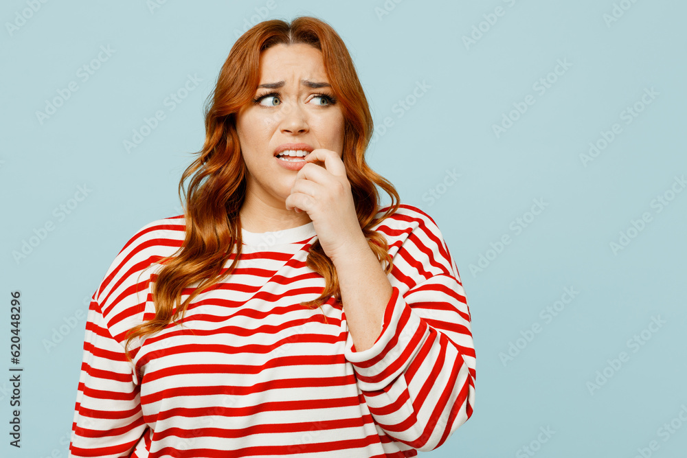 Young sad confused chubby overweight woman she wear striped red shirt casual clothes look aside biting nails fingers isolated on plain pastel light blue background studio portrait. Lifestyle concept. Young sad confused chubby overweight woman she wear striped red shirt casual clothes look aside biting nails fingers isolated on plain pastel light blue background studio portrait. Lifestyle concept.