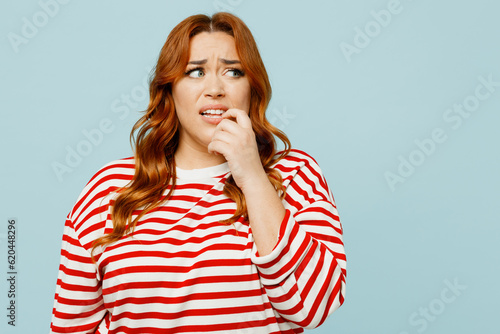 Young sad confused chubby overweight woman she wear striped red shirt casual clothes look aside biting nails fingers isolated on plain pastel light blue background studio portrait. Lifestyle concept.