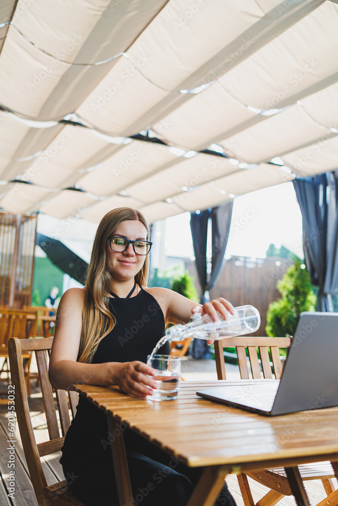 Female manager, freelancer, enjoying coffee break on summer terrace of cozy cafe, working remotely on laptop, smiling looking at laptop. Work online in the summer