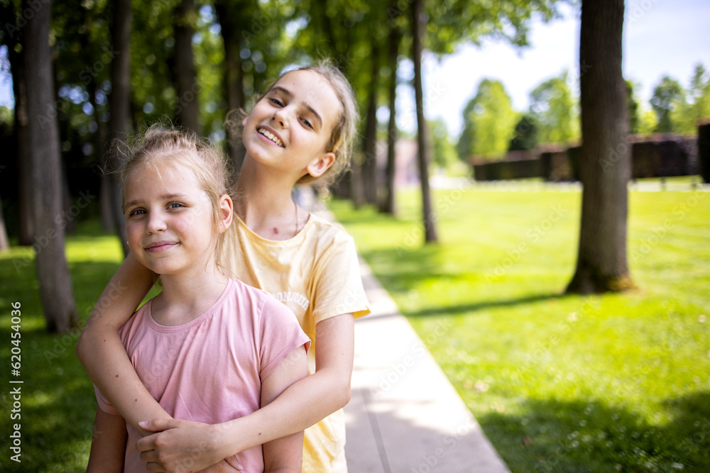 Two girls sisters of school age hug each other. A walk through the summer city park. Love and friendship within the family Two girls sisters of school age hug each other. A walk through the summer city park. Love and friendship within the family