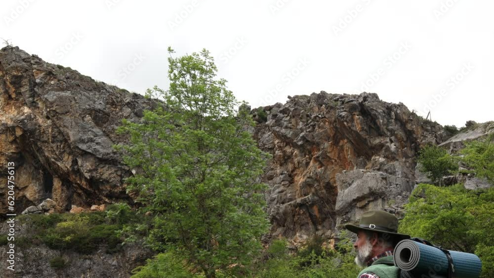 Active man with backpack and hat walking through the rocky meadows at the foot of the mountains Active man with backpack and hat walking through the rocky meadows at the foot of the mountains