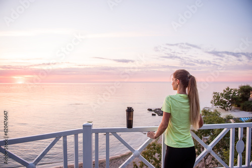 Portrait of young woman in sportswear who stopped for break between workouts. Sportswoman looks at dawn of sun on the sea. Girl walks along the embankment after her morning routine. Outdoor workout