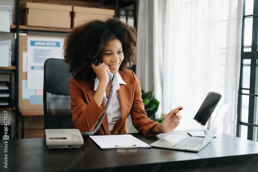 Obraz premium Business African woman Talking on the phone and using a laptop with a smile while sitting at office