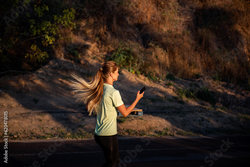 Slender girl in sportswear is jogging, holding smartphone in hands. Young business woman talking on the phone while running on the track in the park. Scattered attention. Striving for goal. Lifestyle