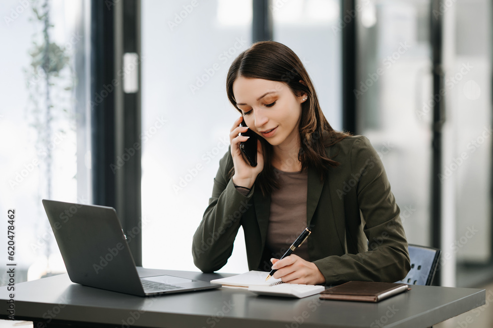 Business Caucasian woman Talking on the phone and using a laptop with a smile while sitting at modern office Business Caucasian woman Talking on the phone and using a laptop with a smile while sitting at modern office