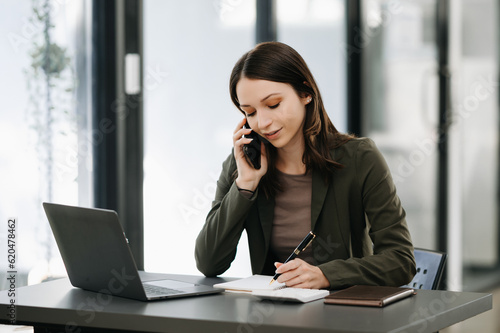 Business Caucasian woman Talking on the phone and using a laptop with a smile while sitting at modern office