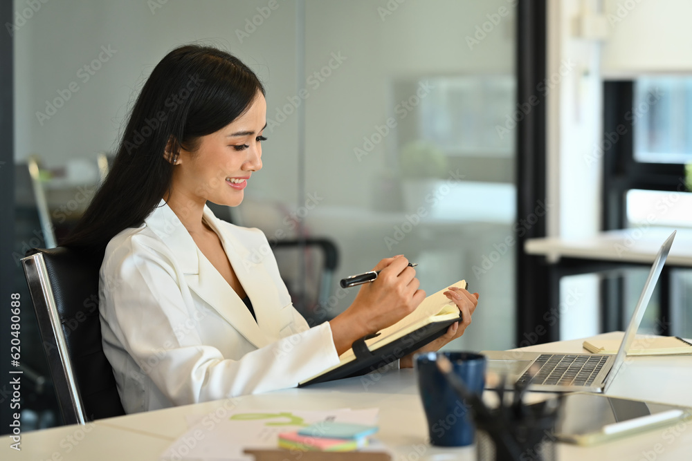 Pleasant businesswoman sitting at working desk and checking schedule or handwriting important information Pleasant businesswoman sitting at working desk and checking schedule or handwriting important information