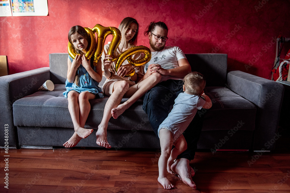 Obraz premium Little girl in festive cap stuck head into the foil baloon ONE. Portrait of happy, diversity family that fools around, having fun for year old baby. Birthday party at home. Lifestyle atmosphere