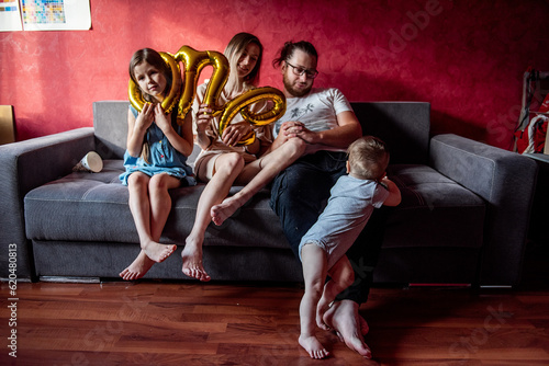 Little girl in festive cap stuck head into the foil baloon ONE. Portrait of happy, diversity family that fools around, having fun for year old baby. Birthday party at home. Lifestyle atmosphere