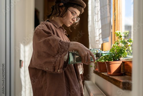Caring creative teenage girl watering houseplant from bottle on wooden windowsill. Home help parents. Hobby growing plant. Sunlight in room warm light. Daughter takes care domestic flowers in pots.