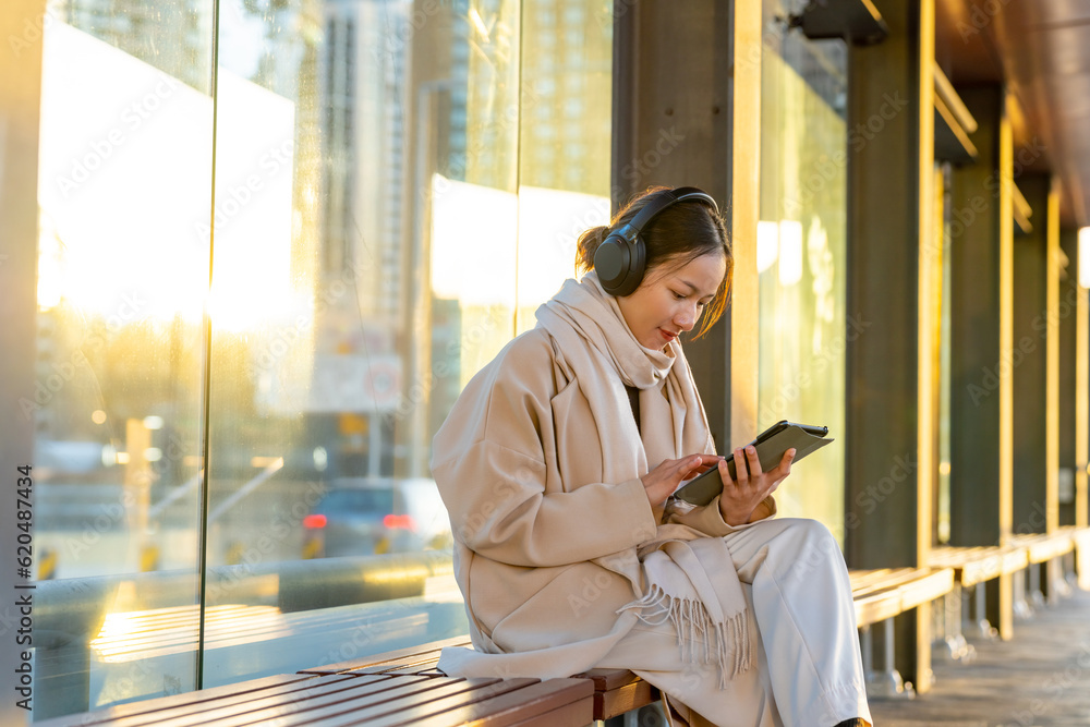Asian woman listening to music from headphones and working on digital tablet during waiting for bus in the city. Attractive girl enjoy urban outdoor lifestyle travel with internet on gadget device.