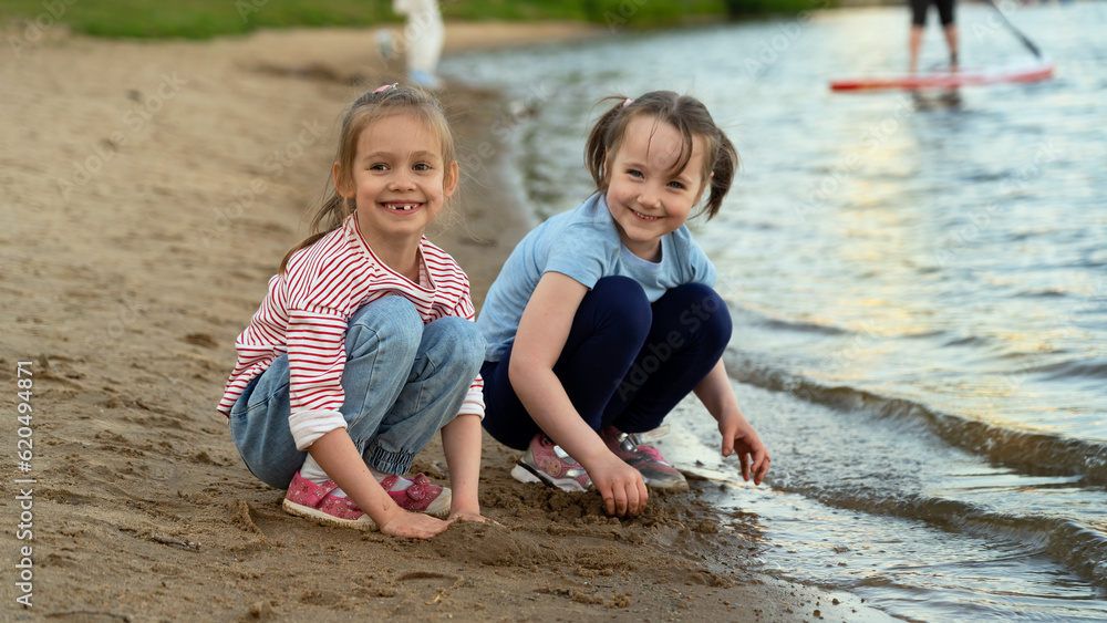 Happy little girls laugh and play with sand on river bank in green summer park at sunset. Active childrens leisure games outdoors by water Happy little girls laugh and play with sand on river bank in green summer park at sunset. Active childrens leisure games outdoors by water