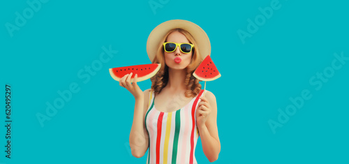 Summer portrait of happy young woman posing with fresh juicy fruits, lollipop or ice cream shaped slice of watermelon wearing straw hat, sunglasses on blue background