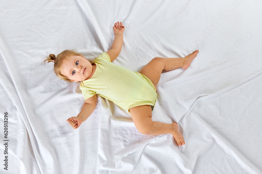 Top view portrait of cute, beautiful, little baby girl, toddler in yellow clothes lying on bed against white studio background Top view portrait of cute, beautiful, little baby girl, toddler in yellow clothes lying on bed against white studio background