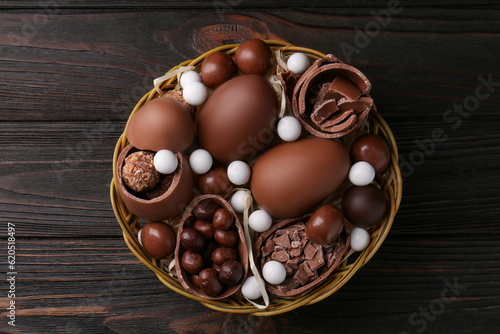 Tasty chocolate eggs and sweets in wicker basket on wooden table, top view