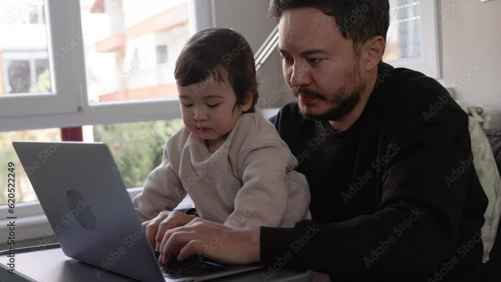 A young man trying to work on laptop holding his son at home. Remote work A young man trying to work on laptop holding his son at home. Remote work