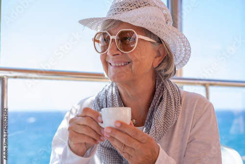 Smiling handsome senior woman sitting inside a cofe enjoying an espresso coffee cup. Elderly woman with hat and sunglasses enjoying summer vacation at sea photo