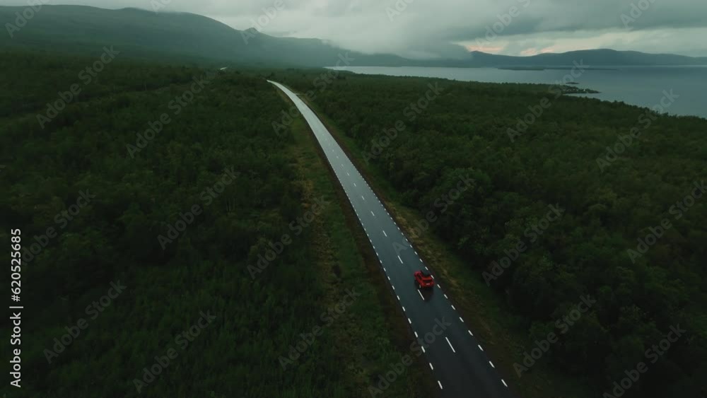 Drone flies over red SUV driving alone on road in the middle of forest with epic nordic fjords and lakes in background Drone flies over red SUV driving alone on road in the middle of forest with epic nordic fjords and lakes in background