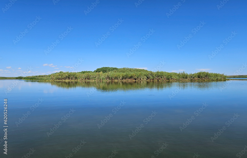 Outdoor summer nature landscape of a small island covered in green grass and reflected in the surface of a still lake under a clear blue sky.