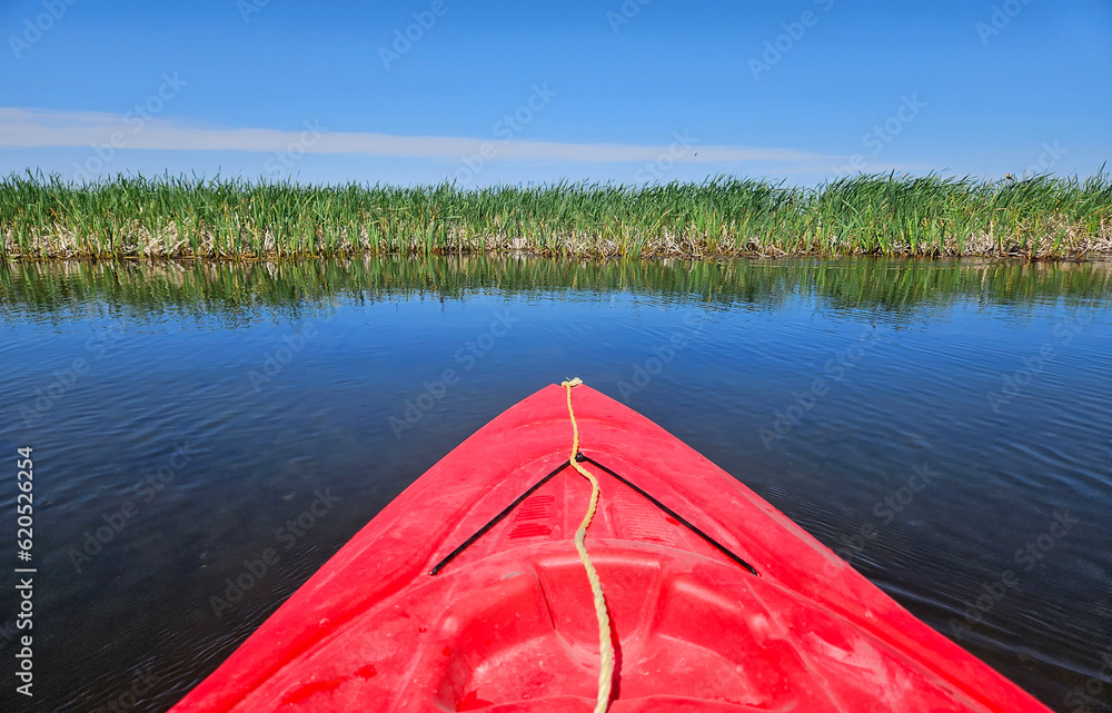 Outdoor recreation image of the bow of a red kayak floating on a lake and pointed toward the grass and reeds along the lakeshore.