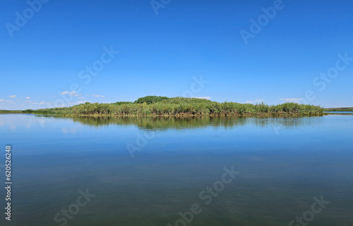 Outdoor summer nature landscape of a small island covered in green grass and reflected in the surface of a still lake under a clear blue sky.
