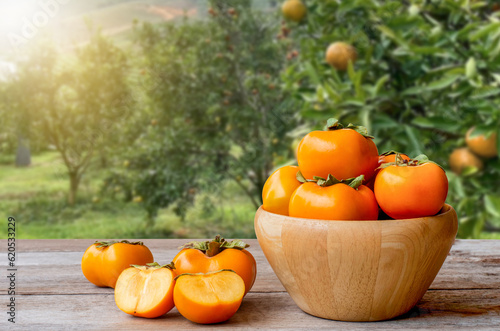 Persimmons or Persimon fruits in wooden bowl on old wooden table with persimmon tree plantation background. photo
