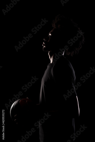 Side lit silhouette of a basketball player. African American man holding basket ball posing against black background.