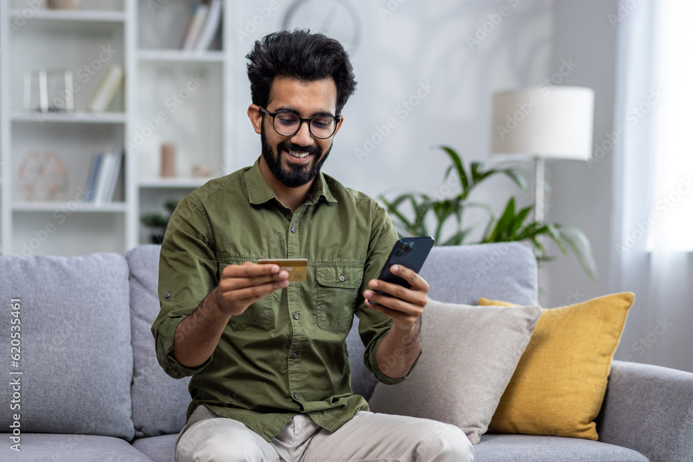 Happy and satisfied man shopping online sitting on sofa at home, hispanic customer holding bank credit card and phone, using online shopping app.