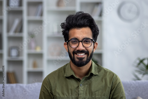 Photos Close-up portrait of young smiling hispanic man smiling and looking at camera while sitting on sofa in living room