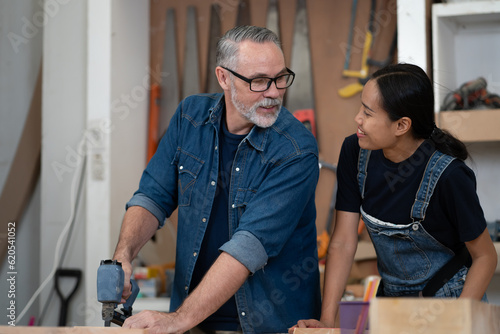 Happy multicultural family working together at wood carpenter shop as owner. Portrait, Asian wife standing with her carpentry husband in furniture business. Diversity ethnic couple in artisan woodwork