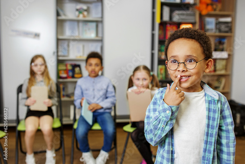 Schoolboy standing in middle of classroom and answering question in front of classmates and teacher.