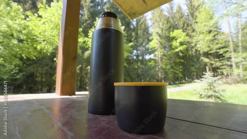 A thermal bottle with a lid for a mug on a table in a forest - closeup A thermal bottle with a lid for a mug on a table in a forest - closeup