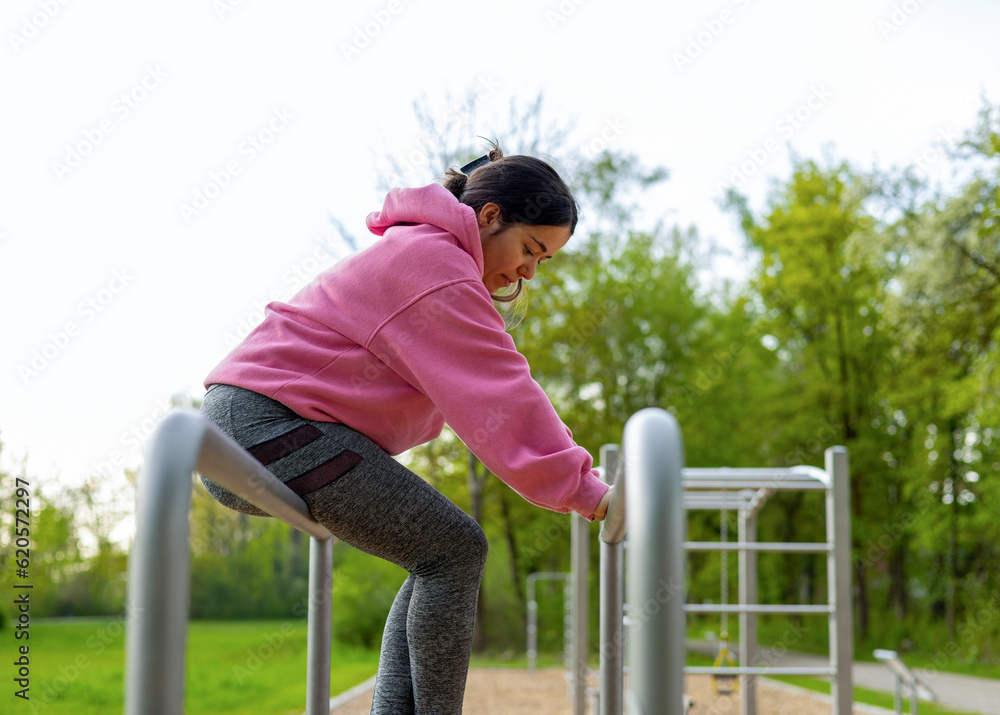 Fototapeta premium young woman in outside outdoor gym using cable chest for workout or parallel bars.girl female is resting sitting down drinking water from plastic bottle.pink hoodie park environment