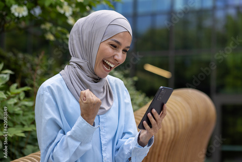 Successful joyful muslim woman in hijab received online win notification, Dinka reading news and holding hand up triumph gesture celebrating sitting on bench outside office building in park photo