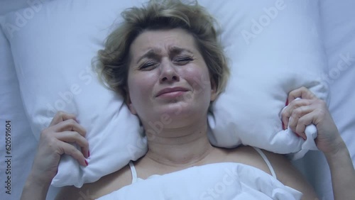 Irritated deeply upset woman adjusting ugly pillow, lying in bed of cheap hotel, has problems with comfort, can't fall asleep because feeling pain in neck and shoulders photo