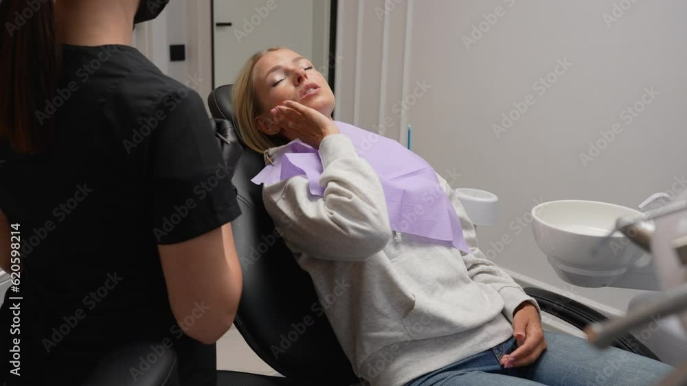 Tracking shot of young blonde woman with toothache lying on dental chair consulting with unrecognizable female dentist and talking during reception in dentistry clinic. Shooting in slow motion. Tracking shot of young blonde woman with toothache lying on dental chair consulting with unrecognizable female dentist and talking during reception in dentistry clinic. Shooting in slow motion.