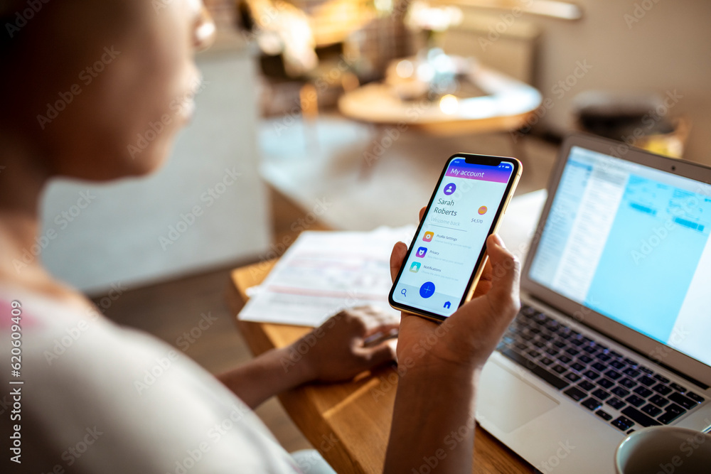 Young woman using a banking app on her phone while working from home on her laptop Young woman using a banking app on her phone while working from home on her laptop