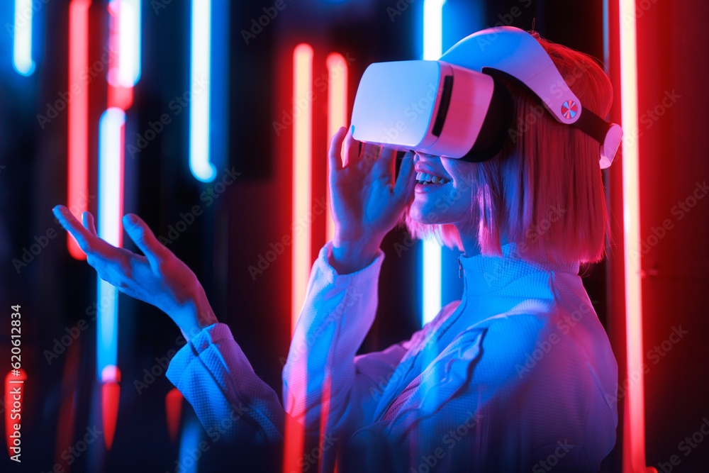 Woman in futuristic costume. Girl in glasses of virtual reality while touching air. Augmented reality game, future technology, AI concept. VR. Neon blue and red light. Dark background. Woman in futuristic costume. Girl in glasses of virtual reality while touching air. Augmented reality game, future technology, AI concept. VR. Neon blue and red light. Dark background.