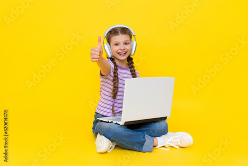 Internet communication for children. A young girl with a laptop in her hands is sitting on the floor and giving a thumbs up. The child communicates online. Yellow isolated background.