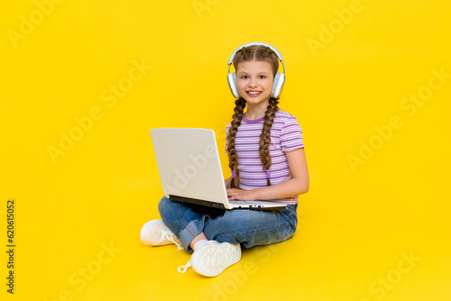 A young girl with a laptop. A little girl is sitting cross-legged on the floor with headphones on, and holding a laptop in her hands. Social networks for teenagers. Yellow isolated background.