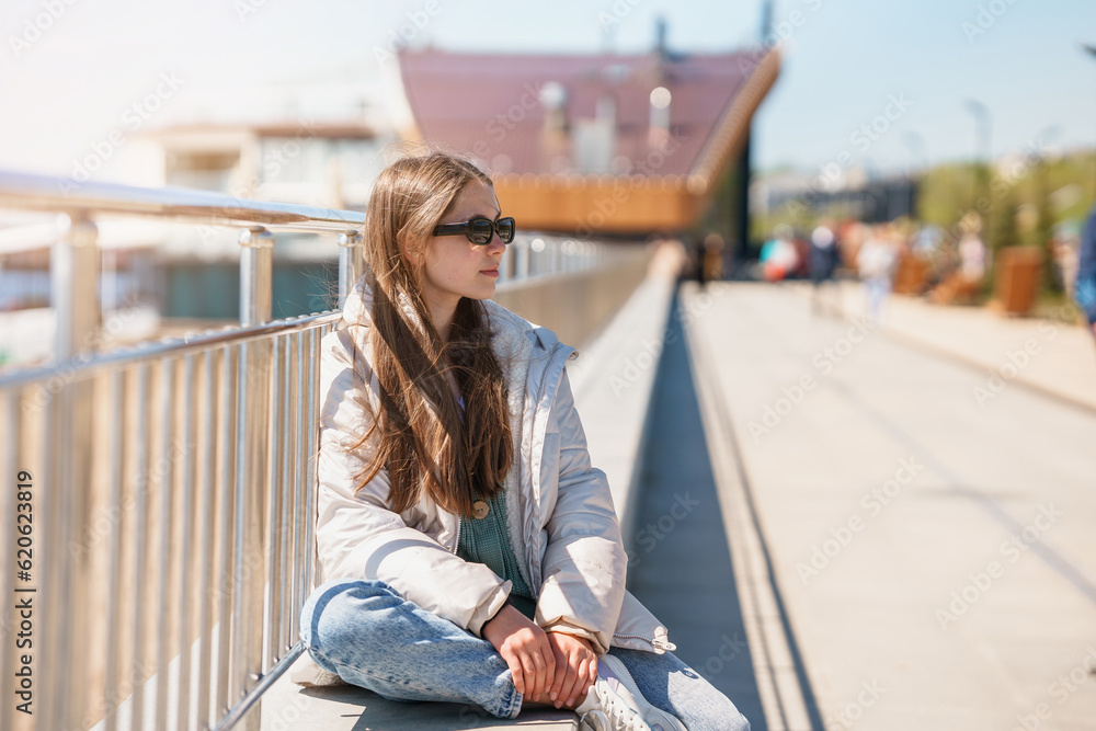A 12-year-old teenager girl in sunglasses is sitting on the embankment by the fence A 12-year-old teenager girl in sunglasses is sitting on the embankment by the fence