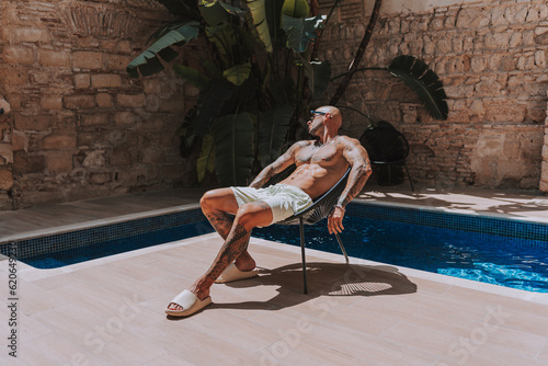 Chico joven musculado y tatuado posando  sin camiseta en piscina de estilo  boho en un hotel un dia soleado de verano photo